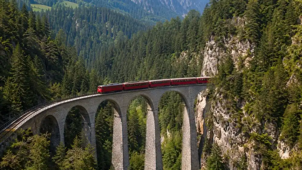 Cerveny uzkorozchodny vlak na kamennem viaduktu nad zalesnenou roklinou u Klosters
