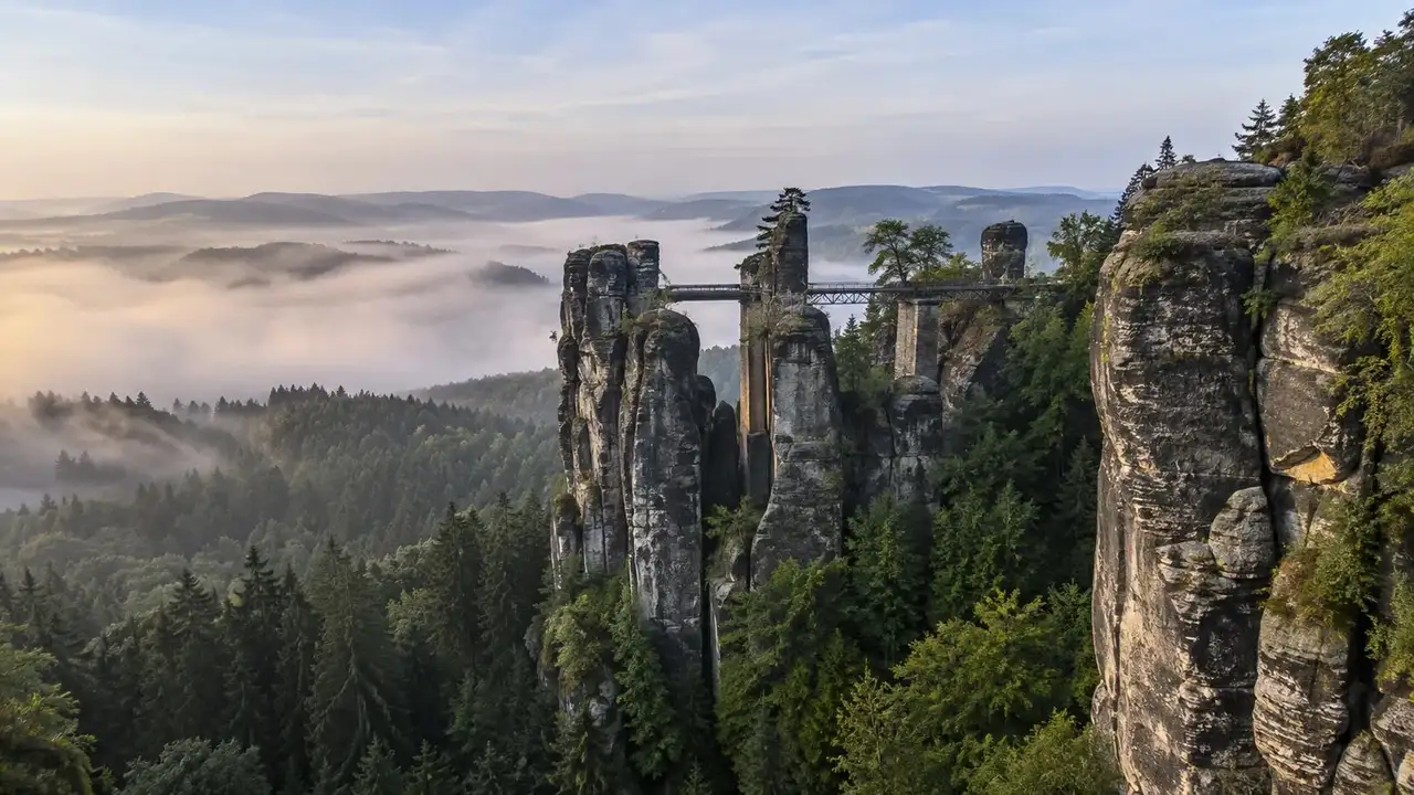 Pískovcové skalní věže s železnou lávkou nad zalesněným údolím v mlze